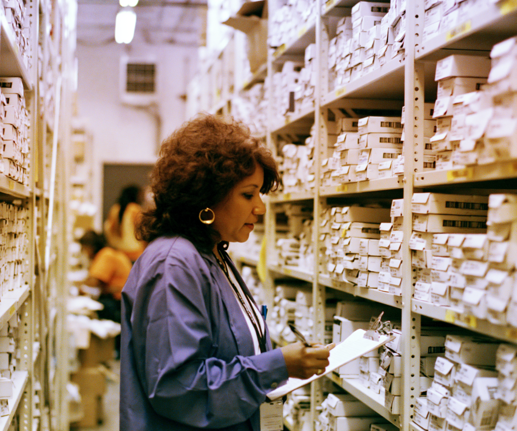 A store owner reviews inventory with a clipboard among stocked shelves in her stockroom