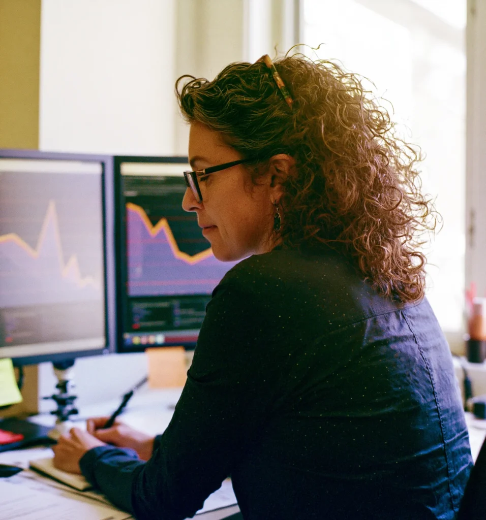 An analyst jots notes at a desk with two monitors showing out-of-focus data dashboards