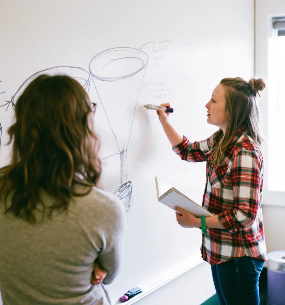 A founder draws a strategy funnel on a whiteboard while a colleague takes notes