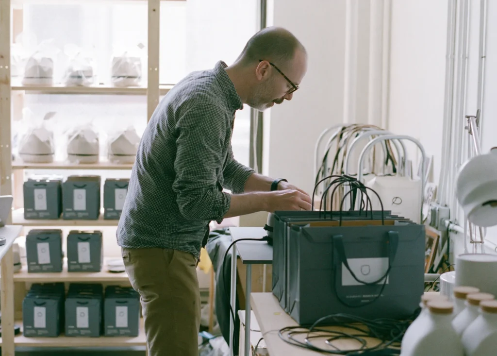 A founder packs branded bags of product at a table in his studio workshop beside shelves of packaged goods