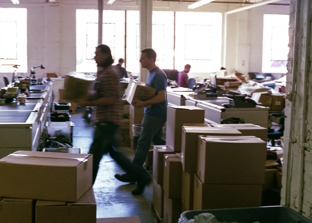 Two ecommerce operators carry boxed orders past packing stations in a bright converted warehouse