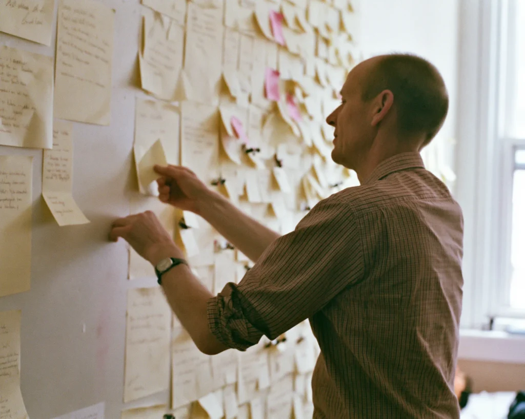A man moves a single sticky note from a crowded idea wall to a nearly empty wall