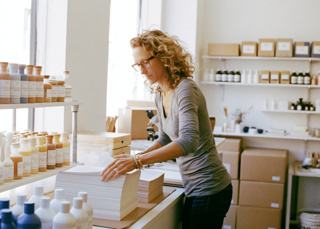 A store owner sorts stacks of printed pages at her studio counter beside shelves of product bottles
