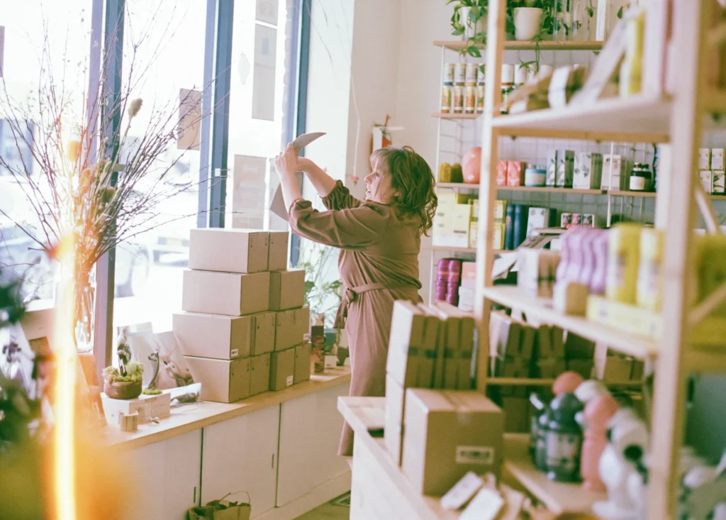 A shop owner hangs signage in her store window while stacked ecommerce shipping boxes wait on the sill