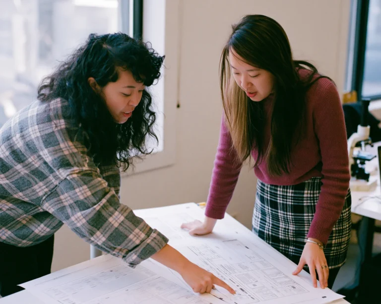 Two specialists review printed website wireframes spread across a worktable