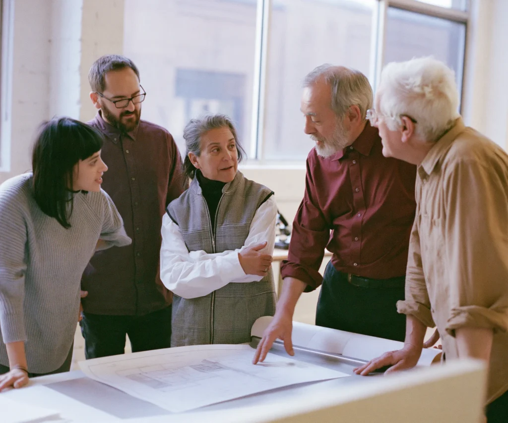 A small team gathers around a single printed plan on a worktable in discussion